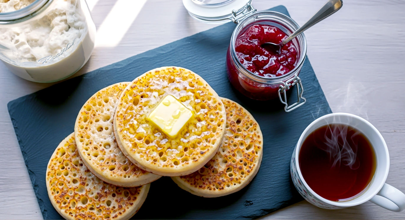 A vibrant, top-down "flat lay" photograph in the style of a modern British food blog. The scene features a stack of freshly made, golden-brown sourdough crumpets on a slate serving plate. One crumpet has a generous knob of butter melting into its porous surface. To the side, there is a small jar of strawberry jam and a cup of tea. In the corner of the frame, a glass jar containing sourdough discard is partially visible, connecting the final product to its origin. The lighting is bright and cheerful, creating an irresistibly delicious and homely atmosphere.