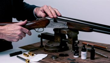 A hyper-realistic, professional photograph in the style of a Fieldsports magazine feature. An experienced gunsmith with weathered hands is meticulously cleaning a classic British side-by-side shotgun. The firearm rests in a dark wooden vise on a rustic workbench, surrounded by an array of bespoke cleaning tools: brass jags, carbon fibre rods, small bottles of oil, and clean white patches. The lighting is soft and warm, coming from a low-angled window, highlighting the rich grain of the walnut stock and the intricate engraving on the receiver. The mood is one of quiet concentration, heritage, and timeless craftsmanship.