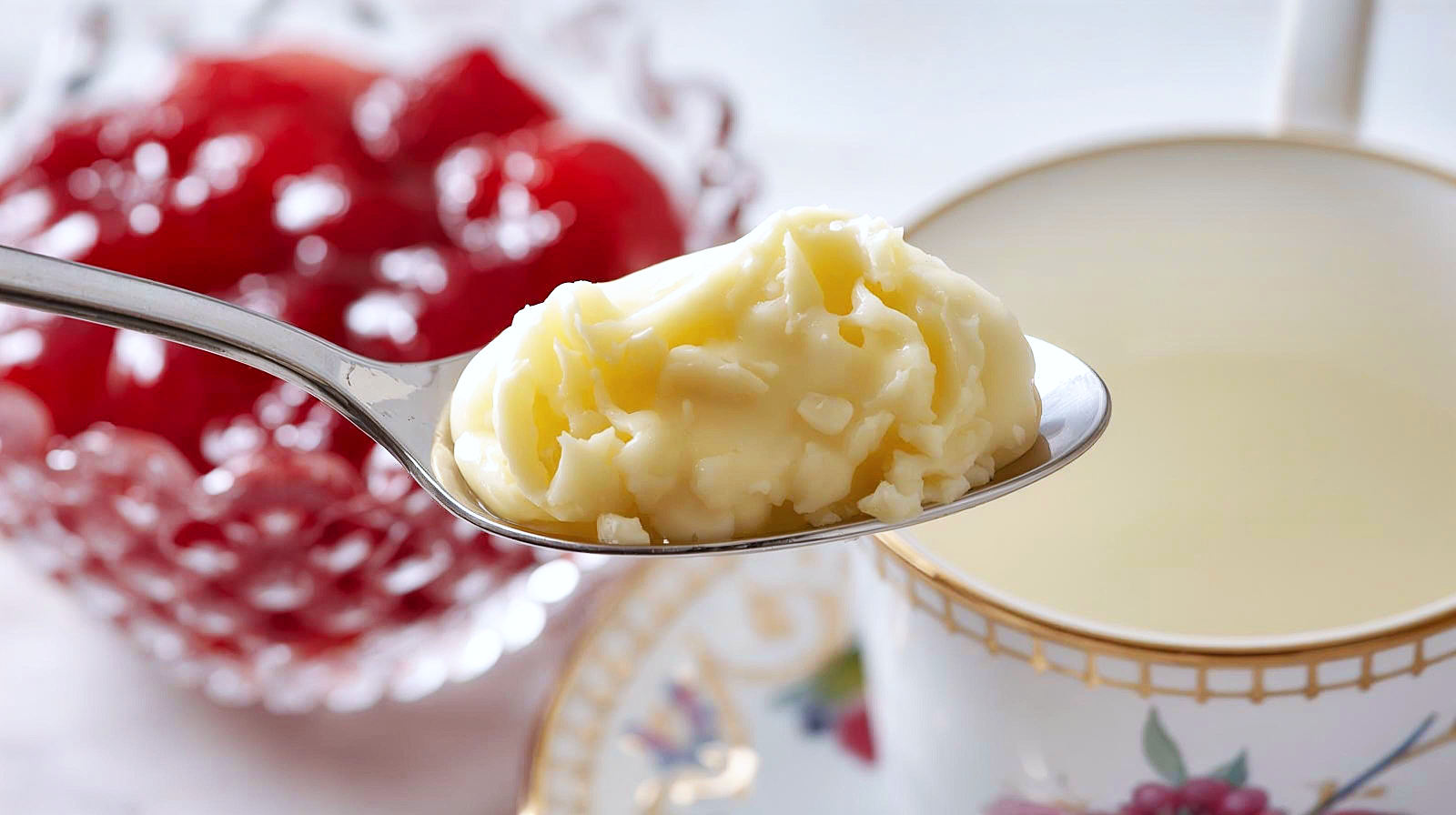 A rich, textural macro shot focusing purely on the toppings.
Focus: Extreme close-up of a silver spoon breaking the golden-yellow "crust" of a pot of authentic Cornish clotted cream. In the immediate background, slightly out of focus, is a crystal dish of glistening, ruby-red strawberry jam with chunks of fruit visible, and the rim of a fine bone china teacup.
Atmosphere: Indulgent and rich.
Lighting: glistening highlights on the jam and the creamy texture of the dairy.