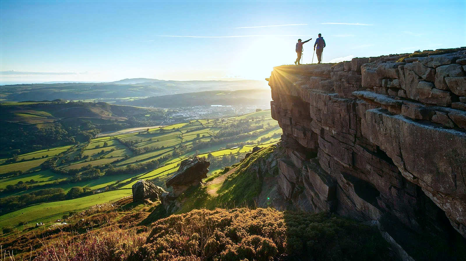 A stunning, wide-angle action shot of two hikers standing atop a dramatic gritstone overhang at Stanage Edge. The camera angle is slightly from behind and above, looking out over the vast, patchwork green valley and distant hills below. The sun is low, casting long, dramatic shadows across the rock face and illuminating the heather in the foreground. The hikers are geared up in practical walking wear, pointing towards the horizon. Style: Adventure travel photography, sharp focus, vivid depth of field, golden hour lighting.